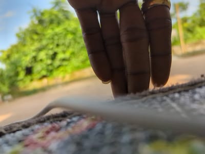 The image features a close-up view of a person's hand, with four fingers visible and extended. A gold-colored ring is worn on one of the fingers. The hand is positioned against a blurred background that suggests an outdoor setting. Lush green foliage and a bright blue sky are discernible, indicating a daytime scene with pleasant weather. The focus is primarily on the texture and details of the hand, with the background serving as context rather than a primary subject. The overall impression is that of a casual moment captured outdoors in a natural environment, possibly in Elele Alimini, Nigeria. There is no visible text in the image.