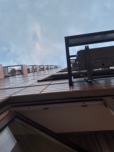 This media file is a vertically oriented, low-angle shot of an apartment building's exterior. The dominant subjects are the building's facade, characterized by vertical siding and a series of balconies, and the cloudy sky above. Several outdoor air conditioning units are mounted on brackets attached to the building's exterior, some with protective grated enclosures. The building appears to be of modern construction, possibly a high-rise, with a repetitive balcony design featuring metal railings and a perforated grate on the outer edge.
The scene is set on the exterior of an apartment building, with a view looking upwards. The time of day is likely daytime, given the illumination, but the presence of clouds suggests overcast conditions, perhaps late afternoon or early morning before the sun is high. The sky is a pale blue with significant cloud cover, indicating a mild or cool day. The image offers no explicit clues to the specific location other than what is provided in the context (Drozhzhino, Russia). There are no people or animals visible in the frame.
A notable detail is the presence of multiple air conditioning units on various levels of the building, suggesting a residential or office complex. The perspective looking up from a low angle emphasizes the height and scale of the structure. The material of the building's siding appears to be a reflective metallic or composite material, catching some diffused light from the sky. The edge of a window frame, with a slightly opened window, is visible on the right side of the frame, suggesting a view from a lower vantage point looking up towards the upper floors. No text is visible in the media file.