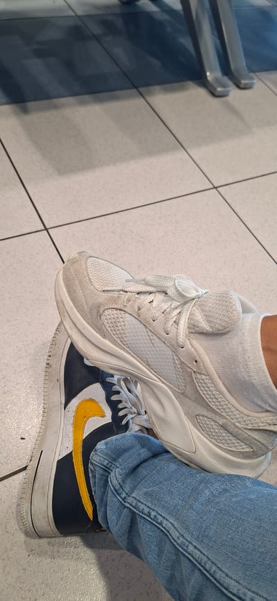This media file shows a close-up of a person's legs and feet, with one foot placed on top of the other. The person is wearing blue jeans. The top foot is clad in a white athletic shoe with white laces and a white sock. The bottom foot is wearing a navy blue and white Nike Air Force 1 sneaker with a yellow swoosh. The shoes are resting on a tiled floor, which appears to be part of an indoor setting, possibly a public space like an airport or shopping mall, given the presence of what look like handrails or a bench in the background. The lighting suggests it is daytime. There is no visible text in the image. The scene is static, with no apparent activity beyond the positioning of the feet. The setting does not provide specific clues to Rhodes, Greece.
