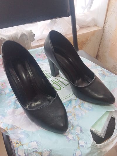 Two black high-heeled shoes are displayed on a table covered with a floral-patterned cloth. The shoes appear to be made of leather. One shoe is positioned on the left, while the other is slightly to the right and behind. The scene is indoors, with a light-colored wall and some white material in the background. The lighting suggests it could be daytime, but the specific time is not discernible. There is some text visible on the insole of the left shoe, appearing to be "5994-2". The overall impression is a simple presentation of footwear, possibly for sale or display. There are no people visible in the image.