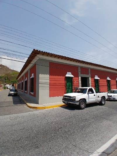 The image depicts a static street scene in San Joaquín, Venezuela, under a clear sky with scattered clouds. A one-story building with red walls, grey trim, and a terracotta tiled roof dominates the right and center of the frame. Its facade features green shuttered windows with white decorative sills and a central green entrance door. A white Chevrolet Silverado pickup truck is parked on the asphalt street, facing left, adjacent to the building's corner. The front end of another white vehicle is partially visible behind the pickup. Further down the street, around a corner to the left, a dark-colored sedan is parked. The asphalt road shows signs of cracking, and a yellow curb separates it from the sidewalk alongside the building. In the distant background to the left, a vegetated hillside is visible. Multiple black utility lines traverse the upper portion of the frame against the sky. No individuals are present in the scene.