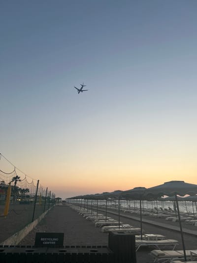 This media file captures a serene beach scene at dusk in Rhodes, Greece. The primary subjects include a row of empty beach chairs and umbrellas lining a sandy path, hinting at a day of relaxation that has concluded. In the sky, a silhouetted airplane is visible, appearing to be in its ascent or descent, adding a sense of travel and transition to the tranquil setting. The scene is bathed in the warm, soft light of sunset, with a gradient of colors from pale blue to a gentle yellow and orange on the horizon.
The overall atmosphere is peaceful and quiet, suggesting a calm evening. The location is identifiable as a beach, likely a resort area, given the organized arrangement of beach amenities. The gentle waves of the sea are visible in the background, further confirming the coastal setting. A volleyball net is also present, implying recreational activities might take place there during the day.
A notable detail is the "RECYCLING CENTRE" sign visible on a dark structure on the right side of the image. The weather appears clear and pleasant, characteristic of an evening in a Mediterranean climate. The time of day is clearly sunset. No people are visible in the frame, emphasizing the quiet and end-of-day feel. The composition creates a sense of vastness with the open sky and the expansive beach, contrasted with the linear arrangement of the beach furniture.