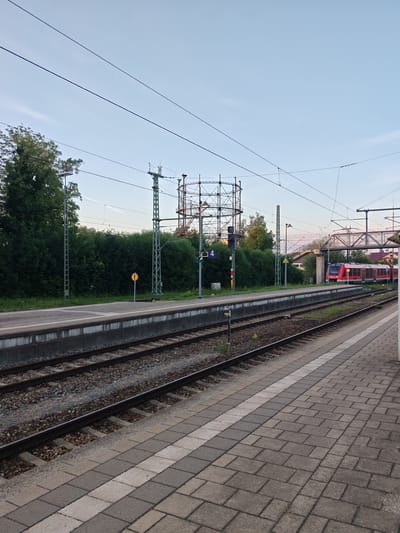The image shows a train station platform in Memmingen, Germany. A red and silver train is stopped on the tracks. There are overhead power lines and a tall metal structure, possibly part of a gasometer, in the background. The platform is paved with grey and white tiles, and there are train tracks running alongside it. Trees and bushes line the edge of the platform. The sky is a pale blue with some clouds.
