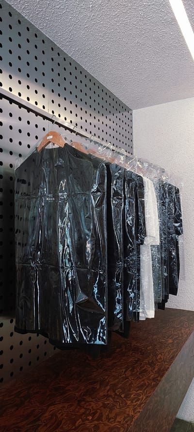 A row of dark garments, individually encased in clear plastic garment bags, hang from a metal rack. The bags are wrinkled, and the garments appear to be shirts or jackets. The first garment from the left has white text on it that reads "AGAIN". To the right of the dark garments, a few lighter colored garments, possibly white shirts, are also visible, similarly bagged. The rack is mounted against a perforated metal wall, which is a dark grey color with evenly spaced circular holes. Above the metal wall is a textured white ceiling. The garments are displayed on a dark brown, polished surface that resembles wood grain. The scene appears to be an indoor retail or storage space, potentially in a clothing store located in Lengkong Kulon, Indonesia.