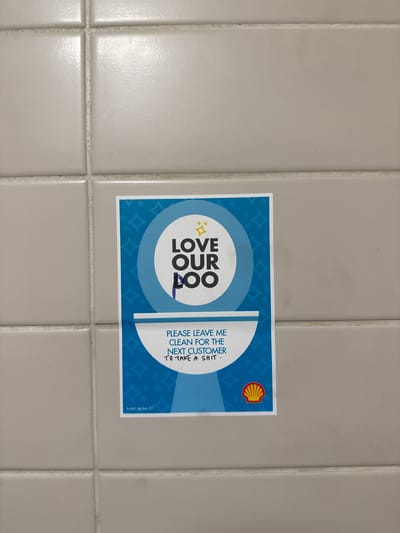 The image shows a rectangular sign affixed to a tiled wall, likely in a restroom. The sign is white with a blue border and a pattern of small diamonds. On the left side, there is a circular graphic with the words "LOVE OUR POO" stacked vertically, with stars above and below the word "OUR". A blue line is drawn on the sign, under the word "OUR". To the right of the circular graphic, text reads "PLEASE LEAVE ME CLEAN FOR THE NEXT CUSTOMER TO TAKE A SHIT." In the top right corner is the Shell Oil logo. The tiles are light gray with darker grout lines. The bottom right corner has the words "toilet.sticker.01". The image does not provide information regarding the location in the city of Derby, United Kingdom.
