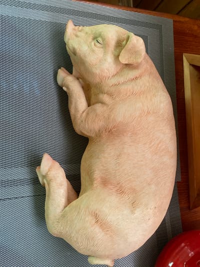 A light pink, ceramic pig figurine is lying on its side on a dark grey placemat. The pig is depicted with its eyes closed, appearing to be sleeping. The figurine has realistic detailing, with textured skin and distinct features like ears, a snout, and hooves. The background shows a wooden surface, likely a table, and a hint of a wooden frame. To the right of the pig, a portion of a shiny, red object, possibly a bowl or decorative item, is visible. The scene is indoors and appears to be a still life arrangement. There is no visible text in the image.
