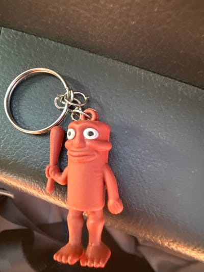 This media file features a close-up, horizontally oriented shot of a small, orange, plastic keychain figure with large, googly eyes. The figure has a simplified, almost cartoonish anatomy with stumpy limbs and a baseball bat held in one hand. It appears to be resting on a dark, textured surface that resembles leather upholstery, possibly from a car seat. A silver keyring is attached to the figure's head, indicating its function as a keychain. The lighting suggests it is daytime, with a warm light source casting a glow on the right side of the image. The background is blurred, but hints at a darker, possibly fabric material. There are no visible text elements in the image. The overall scene is still and inanimate, with the focus solely on the novelty keychain.
