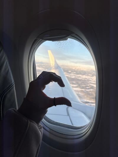 The image depicts an interior view from an aircraft, with an implied location above Teigebyen, Norway. A human right hand, adorned with a ring on the ring finger and a dark band on the pinky, is positioned in the foreground, forming a 'half-heart' gesture. The hand is partially covered by a light-colored, long-sleeved garment with a ribbed cuff. This hand is placed against the oval-shaped airplane window frame. Through the window, a white aircraft wing is visible, extending diagonally from the mid-left towards the lower right. The external environment features a sky gradient from light blue at the top to yellow and orange hues near the horizon, indicative of either sunrise or sunset. Below this, a dense, textured layer of clouds or snow-covered terrain is visible. The upper right portion of the windowpane exhibits small, crystalline formations, likely ice or condensation. A dark, upholstered airplane seatback is partially visible on the left side of the frame.