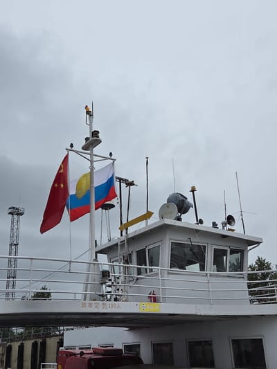 The image captures the upper deck and bridge of a white vessel, likely a passenger ferry, under an overcast sky. The photograph is oriented vertically, showing the ship as if it were on its side.

Prominently displayed from the ship's mast are three flags. At the top is the national flag of Russia, featuring horizontal white, blue, and red stripes. Below it is the flag of the Jewish Autonomous Oblast (J.A.O.), a yellow flag with a central blue horizontal stripe and several narrow rainbow-colored stripes above and below it. The lowest flag is the national flag of China, red with five yellow stars. The flags appear somewhat damp, suggesting cloudy or rainy weather.

The ship's bridge is visible with multiple large windows, and its mast supports various communication and navigation equipment, including antennas, a radar dome, and a loudhailer. White railings run along the deck. In the background, trees with green foliage are visible, along with a metallic lattice structure, possibly part of a crane or tower.

Chinese text is visible in two locations on the ship. A yellow sign with a red fire extinguisher icon displays "最高5米" (zuì gāo 5 mǐ), which translates to "maximum height 5 meters," likely indicating a clearance limit. On the side of the ship's cabin, white characters read "旅客定员130人" (lǚ kè dìng yuán 130 rén), meaning "Passenger capacity 130 people."

The scene depicts a vessel at Nizhneleninskoe, Russia, a known border crossing with China, as evidenced by the flying of both national flags and the regional flag of the J.A.O., located in the Russian Far East. The time of day is daytime, under a grey, cloudy sky.