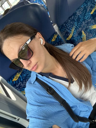 A young adult female is depicted in what appears to be a public transport vehicle, such as a train or bus, situated in the city of Oberndorf, Germany. She is seen from a high angle, reclining against a dark blue upholstered seat with a patterned fabric featuring blue, grey, and yellow abstract designs. Her eyes are obscured by dark-rimmed, rectangular sunglasses with tinted lenses. A white wireless earbud is present in her right ear, along with a small silver hoop earring. She wears a thin silver chain necklace with a small, single-stone pendant. Her attire consists of a light blue, textured, long-sleeved shirt worn over a white shirt with a navy-striped collar. Her long, straight brown hair cascades over her left shoulder, and her right hand is partially visible resting near her collarbone. A black, textured strap with a dark metal buckle, likely from a crossbody bag, is visible draped across her front. The interior of the vehicle shows grey plastic window frames and parts of other seats, while blurred greenery outside the window suggests motion. The subject appears to be resting or asleep.