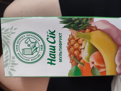 The image shows a close-up of a juice box. The box has a green and white design with an illustration of various fruits, including a pineapple, banana, apple, orange, and peach. The brand name "Наш Сік" (Nash Sik) is prominently displayed in large green lettering. There is also text in a circle that appears to be a logo, with Ukrainian writing. The background of the image is blurred and appears to be a person's shoulder and dark fabric. There is no identifiable information in the image that places it in the city of Krasivka, Ukraine.
