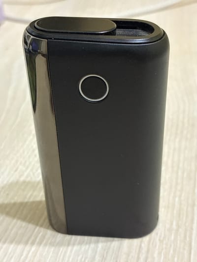 A black electronic device, possibly a portable charger or e-cigarette device, is positioned on a light-colored, textured surface, likely a wooden table or countertop. The device has a rounded top with a small circular button or indicator. A larger, thin circular ring is indented on the front face of the device. The left side of the device appears to have a glossy, reflective black finish, contrasting with the matte black finish of the front and top surfaces. The lighting suggests an indoor environment. There is no visible indication of the city of Melitopol, Ukraine, or any specific urban environment.