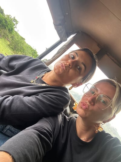 Two women pose inside a vehicle. The woman on the left has dark hair pulled back, a gold necklace, and hoop earrings, pursing her lips. The woman on the right wears gold-rimmed glasses and flower-shaped earrings, also with pursed lips. Both wear dark long-sleeved shirts. The car's interior is visible, and glimpses of green landscape can be seen through the windows.