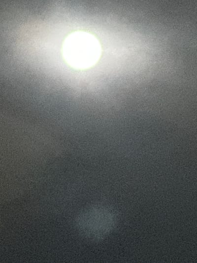 This image is situated in Vevey, Switzerland. The scene is dominated by an intensely bright, circular white light source with a distinct green perimeter, positioned centrally in the upper third of the frame. This light illuminates the immediate surrounding area with a soft, diffuse glow against an otherwise dark, uniformly textured, and heavily granular background. A secondary, significantly fainter, and more diffuse light anomaly, irregular in shape and greyish-white, is present in the lower center portion of the frame. Due to extreme optical blur, extensive image noise, and a profound lack of focus, no discernible structures, human figures, specific objects, or environmental details typically associated with a city setting are visible. The visual data does not permit identification of people, subjects, actions, or interactions within this abstract presentation of light and shadow.