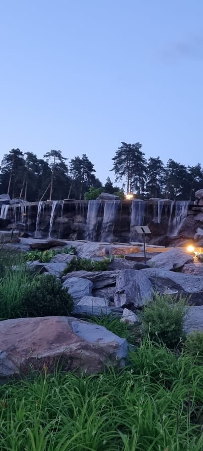 The image depicts a man-made waterfall cascading over rocks, situated in an outdoor environment. Tall pine trees form a dark silhouette against a clear, pale blue sky at dusk. The foreground is filled with large grey and reddish-brown rocks and lush green grass. Artificial lighting is visible on the right side of the scene, illuminating some of the rocks and the waterfall. A small signpost is also present near the waterfall. The scene is serene and appears to be a landscaped area, possibly a park or garden. There are no people visible in the image. The location is identified as being in the city of Shulgino, Russia.