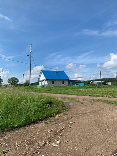 The image depicts a rural scene in Barsai, Russia, under a bright blue sky with scattered white clouds. A dirt road, with visible tire tracks and scattered pebbles, leads into the frame from the foreground, curving slightly to the right. To the left of the road, tall green grass and other vegetation grow.
In the middle ground, a light-colored brick house with a distinctive bright blue, sloped roof stands prominently. The house has a single window visible on its side and a small green fence in front of it. Adjacent to the house, there is a teal-colored shed or outbuilding. Further to the right, another outbuilding with a wooden exterior and a metal roof is visible, along with a wooden fence and a dense line of trees forming the background.
Several utility poles with wires stretch across the sky, indicating the presence of electricity and possibly telephone lines. One pole is located to the left of the house and another to the right, closer to the background. A parked white car is partially visible near the teal outbuilding. The overall environment is natural and appears to be a residential area in the countryside.