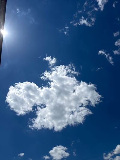 The image depicts a bright, sunny sky over Philadelphia, United States of America. The scene is dominated by a deep blue sky filled with scattered white clouds of varying sizes and densities. A large, fluffy cloud formation is the most prominent feature, positioned in the center-right of the frame. Smaller, wispy clouds are spread throughout the sky, suggesting fair weather conditions.

The sun is visible in the lower-left corner, shining intensely and creating a lens flare that radiates outwards. The bright light indicates it is likely daytime, possibly mid-morning or early afternoon. At the very bottom of the image, a dark, brick-like structure is visible, suggesting the photo was taken from ground level, looking upwards. The presence of power lines or cables can be seen in the upper right quadrant, indicating an urban or suburban environment. A sliver of the crescent moon is faintly visible in the upper center of the frame.

There are no people or animals present in the image. The overall mood conveyed is one of a pleasant, clear day. No text is visible in the media file.