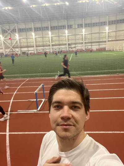 A man in a white shirt is taking a selfie indoors. He's on a red running track with white lane markers. In the background, there's a green soccer field behind a net. Several people are on the field. A hurdle is also visible on the track. The building has a high ceiling with lights and beams.