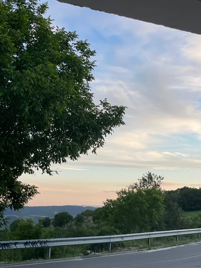 The image depicts a scenic view of a rolling landscape under a sky with scattered clouds, suggestive of late afternoon or early evening. A prominent tree with dark green foliage occupies the left side of the frame, its branches extending into the upper portion of the image. Below the tree, a metal guardrail runs horizontally along the edge of an asphalt road, separating it from a vegetated embankment. In the background, hills and valleys covered in lush greenery stretch towards the horizon, with hints of buildings or structures visible amidst the trees. The sky transitions from pale blue near the horizon to a lighter, cloud-filled expanse above.
The specific location is identified as Valkosel, Bulgaria, known for its picturesque natural surroundings. The scene captures a serene moment, characteristic of rural Bulgarian scenery. There are no people or animals visible in the image, nor any discernable actions or interactions taking place. The focus is on the natural environment and the road infrastructure.