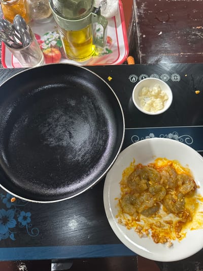 A black frying pan sits next to a white dish of shrimp coated in sauce. Adjacent to the pan, there is a small white bowl filled with chopped garlic. A tray with multiple glass jars containing various ingredients and utensils is located to the left of the pan. The items are arranged on a dark countertop or stovetop.