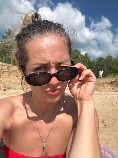 A medium shot captures a woman in the foreground, sitting on a sandy beach under a bright, cloudy blue sky. She is looking directly at the camera with a playful, squinting expression, her lips pursed. Her hair is pulled up into a messy bun, with loose strands framing her face. She wears black, oval-shaped sunglasses, which she is adjusting with her right hand, bringing them down slightly on her nose. A silver necklace with a small cross pendant is visible around her neck. She is wearing a red garment, likely a bikini top, and her bare shoulders and upper chest are exposed, showing some sun-kissed skin. The background depicts a sandy bank with dry grass and sparse trees. In the distance, another person, a man wearing a white t-shirt and shorts, is visible standing on the bank, seemingly engaged in an activity. The overall scene suggests a casual day at the beach. The location is identified as the city of Kzyl-Bairak, Russia.