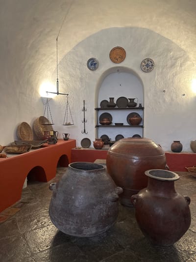 This image captures the interior of a traditional space, likely a historical kitchen or storage area, in Tepotzotlán, Mexico. The scene features a rich collection of antique pottery and household artifacts, displayed against textured white, arched walls and a vaulted ceiling.
In the foreground and to the right, several large, traditional clay pots are prominently featured. Two are reddish-brown, appearing well-maintained, one with a distinctive lid. A third, darker gray pot exhibits a more weathered, matte texture. These substantial vessels rest on a dark, uneven floor and alongside a reddish-orange, low-set counter or shelf.
The white wall in the background incorporates an arched niche, housing wooden shelves that display an array of smaller, dark-colored clay pots, pitchers, and plates. Decorative ceramic plates, adorned with blue and brown patterns, are mounted on the flat wall surfaces, adding splashes of color. To the left, a traditional hanging balance scale, with chains suspending two woven baskets, evokes a sense of historical daily life. Nearby, a wrought iron rack with multiple hooks is also visible.
The space is illuminated by artificial light, casting soft shadows that emphasize the texture of the white stucco walls and the graceful arch of the ceiling. The overall impression is one of cultural preservation, showcasing traditional Mexican craftsmanship and rustic aesthetics. No people are present, and no text is visible in the frame.