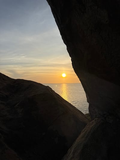 The image depicts a dramatic sunset viewed through a natural rock archway. The foreground is dominated by dark, textured rock formations that create a frame for the scene beyond. These rocks are silhouetted against the vibrant sky and the shimmering ocean.

The main subject is the sun, a bright, warm orb positioned in the upper center of the frame, casting a golden reflection on the rippling water. The sky is a gradient of soft yellows and oranges, transitioning to a lighter blue towards the left. Wispy clouds are scattered across the sky, catching the sunset's light. The ocean is calm, with a metallic sheen from the setting sun.

The activity is simply observing the natural beauty of the sunset. The time of day is clearly sunset, indicated by the low position of the sun and the warm hues of the sky. The weather appears to be clear, with the exception of the thin clouds. The location cues, like the rugged rock formations and the ocean, suggest a coastal environment.

The overall mood is serene and awe-inspiring, evoked by the striking colors and the natural framing of the landscape. There is no visible text in the image. The composition, with the dark rocks contrasting with the bright sky and sea, creates a sense of depth and draws the viewer's eye towards the radiant sunset. The angle of the image suggests it may have been taken from within a cave or a cove.