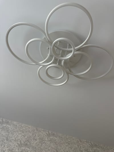 A white, modern ceiling light fixture hangs from a white ceiling. The fixture consists of several intertwined rings of varying sizes, creating an abstract, sculptural design. The rings are all the same white color and appear to be made of a smooth, matte material. The light fixture is the main subject of the image, centered in the upper portion of the frame. Below the ceiling, a patterned wallpaper is visible, suggesting this is an interior shot, likely a room in a residential or commercial building. The overall impression is one of contemporary interior design. The image does not provide any specific location details within the city of Мариинский, Russia.