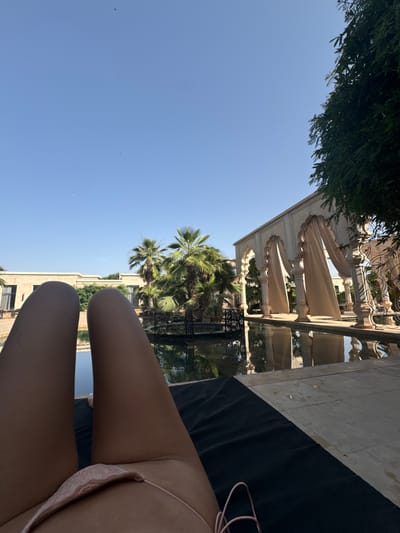 The image shows a person's legs and lower torso in a pink bikini, lying on a black mat. The person is facing towards the camera. The background shows a large rectangular pool of water with the reflection of the sky and the surrounding building. The pool is surrounded by a paved area with trees and an ornate white building structure with archways on the right. The sky is blue with a few white clouds and there are two birds flying in the sky. The location of this image is a resort in the city of Ouled Jelal, Morocco, at latitude 31.664 and longitude -7.87.