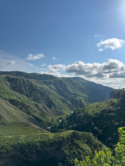 This is a landscape photograph taken near the city of Garni, Armenia. It depicts a steep, green mountain slope under a partly cloudy sky. The mountain is covered in vegetation, with some rocky outcrops visible. The sky is blue with scattered white clouds, and the sun is shining, casting shadows on the mountain. There are no people visible in the image.