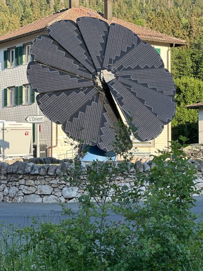 The image shows a close-up of a large, flower-shaped solar panel installation in Le Brassus, Switzerland. The solar panel is composed of many individual segments arranged in a radial pattern, resembling petals. Each segment is dark gray with numerous thin, parallel white lines, indicating solar cells. The solar panel is mounted on a blue base and stands prominently in the foreground.

Behind the solar panel, a stone wall runs horizontally across the middle ground. Beyond the wall, there is a street and a building with a light-colored facade and a red tiled roof. Several windows with green shutters are visible on the building. A street sign pointing to "L'Orient" is also visible on a pole near the building. To the left of the solar panel, a white trailer or mobile unit with a red logo and text "25 EDITION" is parked. In the background, a dense forest of tall green trees covers the hillside. The foreground is filled with lush green foliage, including bushes and grass. The scene appears to be a sunny day.