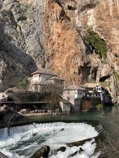 The image displays a complex of traditional stone buildings nestled at the base of a towering rock cliff adjacent to a river featuring a waterfall, located in Dračevice, Bosnia and Herzegovina. A wide, man-made weir in the foreground generates a cascade of white, turbulent water on a turquoise-green river. Several dark boulders are visible within the rushing water. The buildings, constructed primarily from light-colored stone with multi-layered dark stone roofs, exhibit a distinctive architectural style. A prominent multi-story structure features an elevated section with multiple small windows. Directly above the river, a covered outdoor seating area, supported by columns, accommodates approximately ten individuals seated at tables. Additional stone and white-facade buildings line the riverbank to the right. The background is dominated by a massive, stratified limestone cliff, displaying hues from grey to reddish-brown, with scattered green vegetation and a visible cave opening in its upper right section. The scene is illuminated by direct sunlight, casting distinct shadows.