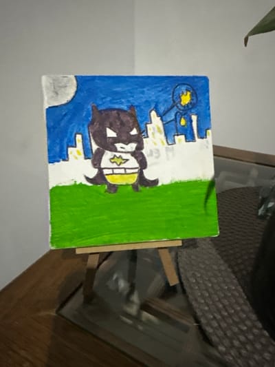 The image displays a small, rectangular canvas painting positioned on a miniature wooden easel. The painting, rendered in a simplistic, childlike style, depicts a stylized, dark-colored Batman figure with a yellow belt and bat symbol standing in the foreground on a vibrant green field. In the background, a blue night sky features a light grey crescent moon in the upper left and illuminated white and yellow outlines of city buildings. To the upper right, a yellow bat-signal is projected within a blue circle, emanating light rays downwards. The easel holding the painting rests on a polished, reddish-brown wooden surface, which itself appears to be part of a furniture piece with a dark, reflective glass top visible beneath. To the right of the easel, a dark, textured object, possibly a woven mat or part of a chair, is present. A dark green plant leaf is partially visible in the upper right quadrant of the image. The scene is illuminated by an indoor light source, casting subtle shadows. This indoor setting is located in Juriquilla, Mexico.