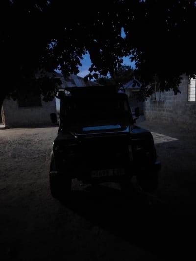 The image displays an outdoor scene in Mwandege, Tanzania, under low light conditions consistent with dusk or dawn. A dark-colored utility vehicle, likely an SUV, is centrally parked on an unpaved, dark ground surface, facing the viewer. The vehicle's front license plate is visible, displaying "T498 EBH". In the background, to the left, a light-colored building with a dark opening is partially visible. To the right, another building constructed of block or stone features a vertically barred window. Overhead, dark tree foliage frames the upper portion of the scene, silhouetted against a pale blue sky. No individuals, discernible actions, or interactions are present.