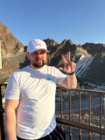 An adult male stands outdoors, facing forward, against a mountainous backdrop in Hatta, United Arab Emirates. He has light skin, a short beard, and is looking directly at the camera. He wears a white baseball cap with a "Paul & Shark Yachting" logo and a white short-sleeved t-shirt featuring a small teal palm tree and sun emblem on the left chest. A dark patterned smartwatch is visible on his left wrist. His left hand is raised, forming a V-sign with his index and middle fingers. He is positioned behind a brown metal railing, likely on an elevated platform or balcony. In the background, rugged, rocky mountains rise under a clear blue sky. To the right, a large concrete structure, identifiable as part of the Hatta Dam, descends the mountainside, featuring prominent murals or artwork. A road is visible at the base of the mountains below the railing, with a white vehicle partially visible near the dam structure. The scene is illuminated by natural sunlight.