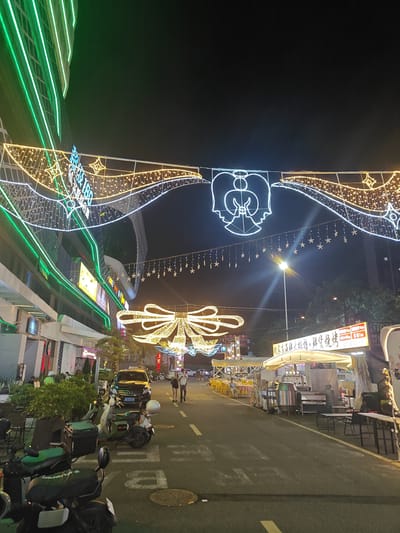 The image captures a street scene at night in Nanhai District, China, illuminated by festive decorations and commercial lighting. A building with green neon accents dominates the left side of the frame, while the street is adorned with elaborate string lights forming wave patterns, an angel silhouette, and a large bow. Various food stalls and restaurants line the right side of the street, some with outdoor seating and yellow chairs. Several parked scooters and a black car are visible on the street. A few individuals are walking or standing in the scene, suggesting a lively evening atmosphere. The overall impression is that of a bustling commercial street during a holiday or special event.