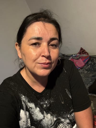 The image is a portrait of a woman in Starokhalilovo, Russia. She is looking towards the camera with her head tilted. The woman has dark hair and is wearing a black t-shirt with a white speckled pattern and a graphic design. She is also wearing hoop earrings. The background is blurred, but it appears to be an indoor setting with a gray wall and some patterned fabric. A small safety pin is visible on her shirt. The lighting suggests it is either daytime or artificial indoor lighting. The woman's expression is neutral.