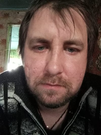 The image is a close-up selfie of a man with light skin, stubble beard, and dark brown hair. He has blue eyes and some facial scarring. He is wearing a black and gray patterned sweater. Behind him, a window is visible, through which bright light is shining. To the right of the window, there's patterned wallpaper. The location is indicated as Parabel, Russia, but no specific details within the city are discernible from the image itself.