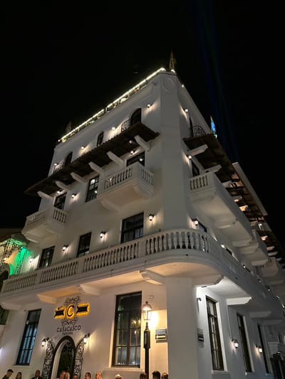 This nighttime image captures the "CASACASCO PANAMA" building, located in Casco Viejo, Panama City, Panama. The multi-story structure features a white facade, illuminated by numerous warm-toned wall-mounted lamps positioned between windows and above balconies. A string of white lights outlines the rooftop perimeter. The building incorporates multiple levels of balconies with white balustrades and dark, possibly wooden, eaves projecting over some sections. A dark flag with a yellow element is visible on the rooftop. The sky above is dark, consistent with nighttime, and a faint blue and green light streak is present in the upper right quadrant, possibly an artifact or light beam. At the ground level, a partially obscured group of individuals is gathered near an arched entranceway beneath the building's signage. A dark street lamp post with a directional arrow sign is positioned to the right of the entrance. To the far left, a section of an adjacent building displays a green illuminated architectural detail.