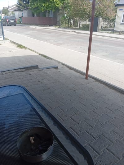 The image shows an outdoor scene in Kostopil, Ukraine. In the foreground, there is a metal ashtray filled with cigarette butts on a black metal table. The table surface has a mesh design. The ground is paved with grey rectangular bricks. A reddish-brown metal pole stands upright near the edge of the sidewalk. A paved sidewalk runs parallel to a street. On the street, a black minivan is driving away from the viewer. Houses and trees line the street. The sky is bright.