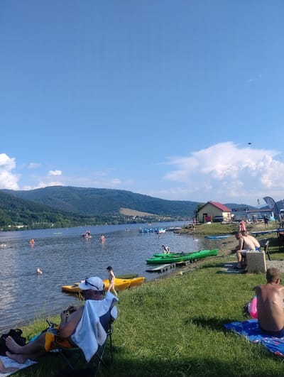 A group of people are enjoying a sunny day by a large body of water, likely Lake Międzybrodzkie, in Międzybrodzie Bialskie, Poland. The water is calm, with several individuals engaged in various water activities, including swimming and paddling kayaks.
In the foreground, two men are seated on a bench near the grassy shore. One is facing away from the camera, wearing swim trunks, while the other is facing slightly towards the camera, also in swim trunks. Further to the right, another shirtless man is sitting on a blue towel on the grass, facing away.
On the lake, numerous kayaks and people on paddleboards are visible. Some are in the water, while others are near the shore. A yellow kayak is positioned on the grass in the middle ground, and several bright green kayaks are lined up by a wooden dock.
In the background, a small building with a red roof is situated on the grassy embankment, adjacent to a larger structure with banners. Rolling, tree-covered hills form the backdrop, meeting a clear blue sky with scattered white clouds. The overall atmosphere is one of leisure and outdoor recreation.