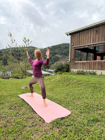 A medium-shot, full-body, eye-level, outdoor photograph shows a woman performing a yoga pose on a pink yoga mat placed on a grassy lawn. The woman has shoulder-length auburn hair and is wearing a long-sleeved, deep purple top and leopard-print leggings. Her arms are raised with palms facing forward, and her body is in a low warrior pose, with her right leg bent and forward. Her left leg is extended backward. In the background, a rustic, wooden building with large windows is visible to the right. Beyond the building and the woman, lush green hills and trees are seen under a cloudy sky, suggesting a rural or mountainous setting, possibly within the vicinity of Ban Khun Wang, Thailand, which is known for its natural beauty and agricultural landscapes. A pair of tan athletic shoes are placed on the grass near the yoga mat to the left.