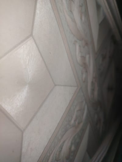 The image shows a close-up, angled view of a textured surface. The surface is divided into geometric panels. One panel on the left has a circular, radiating pattern. Adjacent panels have a fine, parallel line texture. A patterned section on the right features an ornate, light-colored design on a muted green background, resembling a floral or scroll motif. The lighting suggests it is indoors, with shadows indicating a light source from the top right. The overall color palette is neutral, with shades of white, light gray, and pale green. The image is a detailed capture of a decorative surface, potentially wallpaper, ceiling tiles, or a textured wall. The location in the city of Qazax, Uzbekistan cannot be determined from the visual information provided.