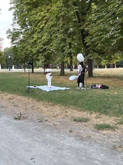 This media file captures a daytime scene in a park setting, likely Parma, Italy, given the context. The primary subjects are two individuals and various photography equipment. One individual, dressed in black, stands near a folding chair with several bags on it, one of which appears to be pink. This person is holding a round, white lighting modifier. Nearby, another individual, wearing a white dress, is posing on the grass, with a long white cloth spread out behind them.
The activity taking place is a photo shoot. The presence of lighting equipment like the softbox and a reflector (held by the person in black) strongly suggests this. The setting is a grassy park with mature trees providing shade. A paved path runs along the right side of the frame. The lighting suggests it's either morning or late afternoon, with the sun casting some shadows. The overall mood seems focused and professional, centered around the creative process of photography. There is no visible text in the image.