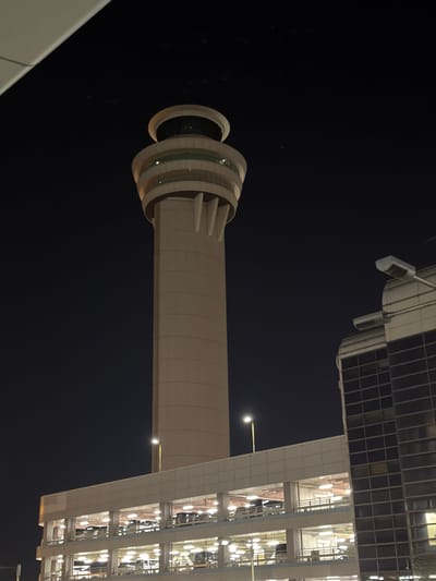 A cylindrical tower with stacked, illuminated rings at its peak stands against a dark night sky. The tower connects to a modern building featuring a grid of windows and illuminated interior spaces. The tower and building architecture are primarily white or light-colored, creating a contrast against the darkness. Streetlights are visible on the building's exterior.