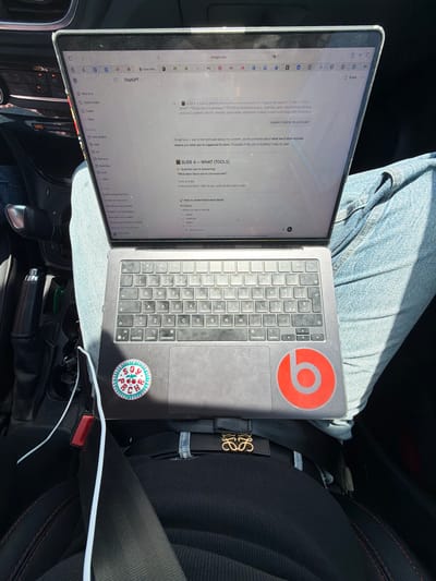 A MacBook laptop rests on a person's lap, covered by blue jeans, inside the passenger seat of a car. The bright sunlight entering from the left indicates it is daytime with sunny weather. The car's interior features black upholstery with red stitching on the seat and gear shifter boot. A black strap with a gold "LOEWE" logo, likely from a bag, is visible secured by the seatbelt. A white charging cable runs along the car's console.
The laptop screen displays a web browser with "chatgpt.com" open, showing a conversation related to a presentation or project. The text "SLIDE 4 - TOOLS (WHAT)" is visible, along with bullet points detailing "PHYSICAL Immersive spaces, materials, water, steam VISUAL Minimal and luxury aesthetic DIGITAL Website, social media, registration HUMAN Guided experience, staff interaction..." The chat interface includes prompts like "explain it better im confused" and a response beginning "Ok got you - you're not confused about the content..." Other visible text includes "New chat," "Q Search chats," "Projects," and a list of names. A warning label on the dashboard reads "SO PASSENGER AIRBAG."
The laptop's lid features a red "Beats" logo sticker, and the palm rest has a round, colorful sticker. The keyboard appears well-used. The setting suggests someone is working or browsing while in transit or parked in Sollana, Spain.