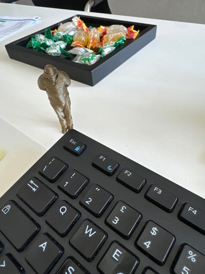 The scene depicts an indoor environment, specifically an office or conference room in Galatina, Italy. A white table surface occupies the majority of the frame. In the bottom right quadrant, a black computer keyboard is partially visible, angled towards the viewer. Notable keys include 'Esc' with a blue padlock icon, 'F1' with a blue crescent moon icon, 'F2', 'F3', '1', '2', '3' (with '£'), and '4' (with '$').
Positioned centrally on the table, between the keyboard and a tray, is a small, bronze-colored humanoid figurine. The figurine appears to be a stylized, armored figure, possibly a character from popular culture, facing away from the keyboard towards the background.
In the upper-middle portion of the table, a black rectangular tray holds an assortment of individually wrapped candies. The candies are primarily in green and yellow/orange wrappers. Identifiable branding on the green wrappers includes "SAILA," and on the yellow/orange wrappers, "Gelées." To the far left, a document with green graphics is partially visible on the white table. A faint glimpse of a person's arm or sleeve is discernable at the extreme top edge of the image.