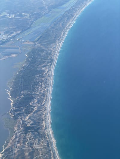 This aerial image offers a panoramic view of the coastal landscape near Grândola, Portugal, under bright, clear daylight. The upper portion of the frame is dominated by the deep blue Atlantic Ocean, where faint lines of white breaking waves define a long, curving sandy beach. A tiny, light-colored speck, possibly a distant boat or aircraft, is visible in the open water.
Immediately inland from the shoreline lies a broad belt of vegetated sand dunes, densely covered with dark green trees, likely pine forests, and scrub. Unpaved roads or tracks cut through these forested areas. Towards the bottom left, the land transitions into a mosaic of large, rectangular agricultural fields, many appearing flooded or cultivated, characteristic of rice paddies or similar water-intensive crops. A small settlement with visible buildings is nestled among these fields.
Further towards the bottom right, an irregular inland body of water, possibly an estuary or coastal lagoon, mirrors the sky. This water feature is also bordered by more organized, rectangular plots and patches of green vegetation. The scene showcases a blend of natural coastal features with significant human impact through agriculture and settlement. No specific human activities are individually discernible, and no text is visible.
