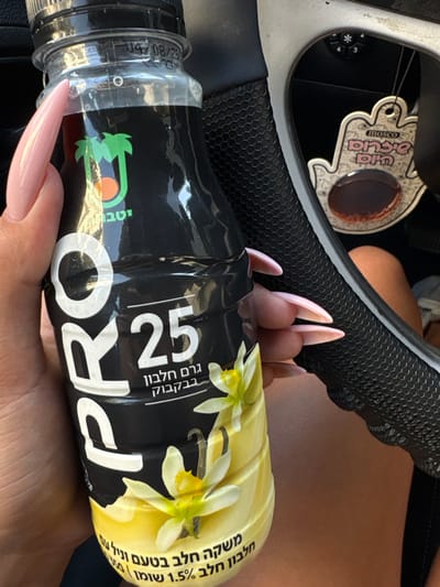 The image shows a close-up of a hand holding a black bottle of "PRO 25" vanilla flavored milk drink. The bottle is being held in the driver's seat of a car. The driver's arm and hand are visible. The bottle has a green palm tree logo with the word "סוגי" written below it, and the number "25" on it. There is a car air freshener in the form of a hamsa (hand) hanging on the driver's side, with a pink background and the word "Mosco" and the Hebrew words "עין הרע" ("evil eye") written on it. The car's steering wheel is visible in the background. The location is Ramla, Israel, as indicated by the coordinates 31.932 latitude, 34.853 longitude.