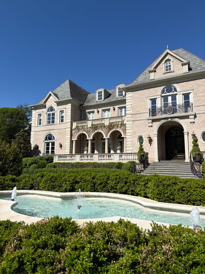 This media file captures a grand, opulent residential estate under a clear, bright blue sky, suggesting a sunny day. The scene depicts a multi-story mansion and an elaborate formal garden with a fountain.
The primary subject is a large building, likely a mansion, constructed with light-colored brick and adorned with numerous stone architectural elements. Its design features strong classical or European influences, characterized by symmetry, arched windows, ornate ironwork balconies, and detailed stone balustrades. Notable architectural details include prominent gabled roofs with dormer windows, some featuring unique quatrefoil-like designs, and others rectangular. The main entrance is particularly grand, with a large arched doorway leading to an impressive black double door, fronted by a wide stone staircase flanked by elegant black wrought-iron railings. Below, multiple arched porticos and additional balustrades add to the building's stately appearance.
Running parallel to the building is a long, tiered stone fountain with light blue water actively flowing and splashing. The fountain is bordered by meticulously manicured, dense green hedges, which also line parts of the building and property, indicating formal landscaping. The overall impression is one of luxury and meticulous maintenance.
No people are visible, lending a serene and quiet atmosphere to the expansive property. The strong sunlight and lack of clouds indicate clear weather, likely midday or early afternoon. The location context of Potomac, United States of America, aligns with the style and scale of this upscale residence. No discernible text is visible within the image.